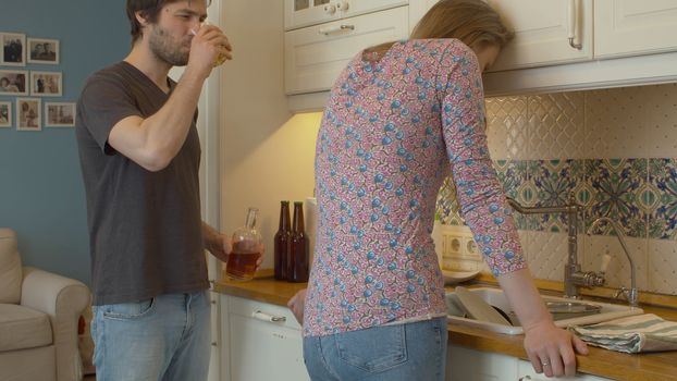 A tired woman in depression standing next to a sink in the kitchen. Alcoholic husband drinking wiskey. Domestic conflict and violence