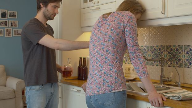 A tired woman in depression standing next to a sink in the kitchen. Alcoholic husband offers her a drink. Domestic conflict.