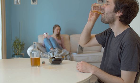 Young man drinking whiskey. In the background, in defocus, the wife sits on the couch and looks at the computer.