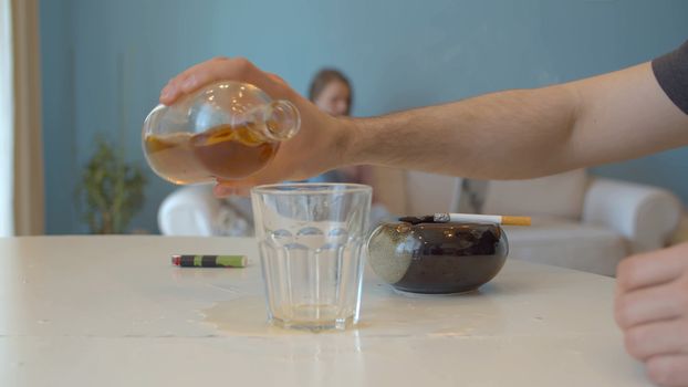 Close up hand of a drunk man pouring whiskey into a glass. On the background, in defocus, the wife sits on the couch and looks at the computer