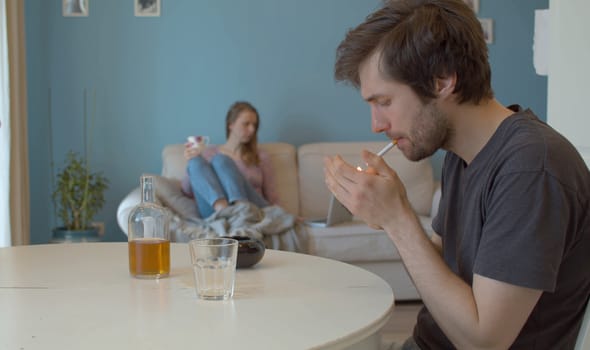 A drunk, sad man lighting a cigarette. In the background, in defocus, the wife sits on the couch and looks at the computer.