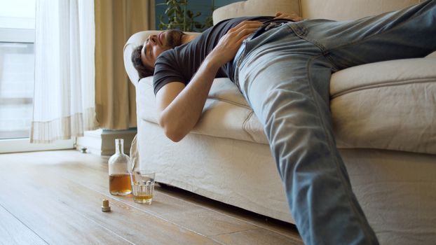 Drunk man sleeping on the couch dressed. Nearby is an unfinished bottle of whiskey and a glass. Young debauchee in his apartment after party