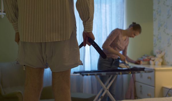 A man in shorts and a shirt holding a belt in his hands waiting while his wife ironing trousers for him. Dominant aggressive husband, domestic violence