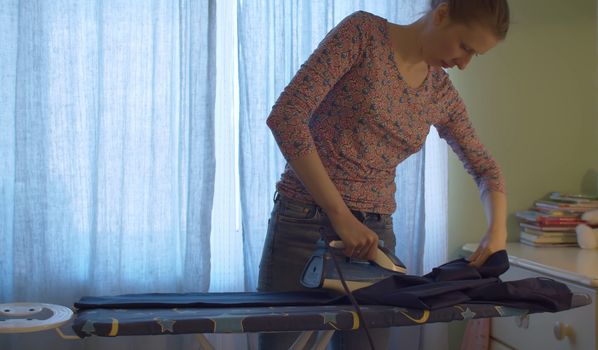 Attractive smiling woman ironing trousers for her husband in bright room. Domestic daily life. Housekeeping