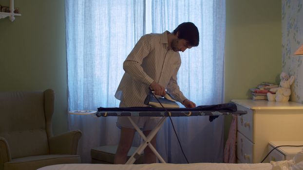 Man in shorts and shirt ironing trousers in bright room. Domestic daily life. Housekeeping man