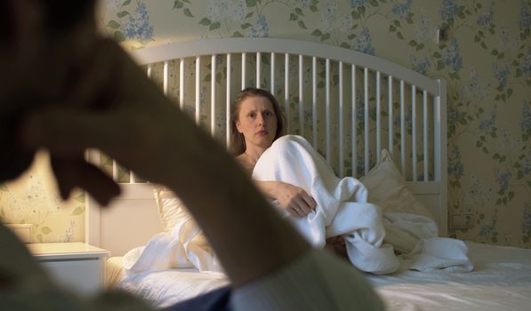 Young beautiful woman in a bedroom. She is alarmed and unhappy. In the foreground a husband waiting for her answer. Sexual harassment in the family