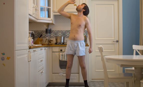 A man in shorts and socks drinking whiskey from a bottleneck in the kitchen. Naked hangover boozer in the morning after the party