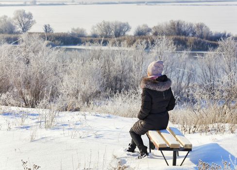 A young woman in the rays of the winter rising sun greets the dawn on the banks of the Southern Bug River, sits on a wooden bench and looks into the distance.