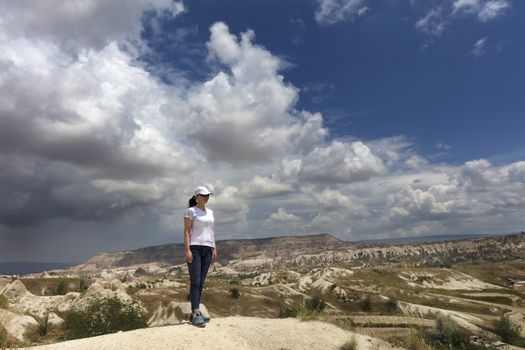A young girl in a white cap and a T-shirt, blue jeans stands on top of a rock in Cappadocia and looks into the distance against the backdrop of a cloudy sky and mountain scenery.