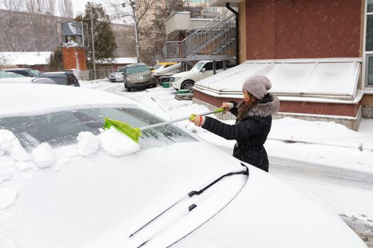 Young beautiful woman in a black coat cleans the snow with a folding green long brush from the surface of her car