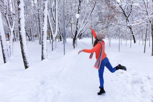 A young beautiful woman in a bright coral jacket and blue jeans in winter walks along the alley of the snow-covered fairy-tale park and throws a large snowball.