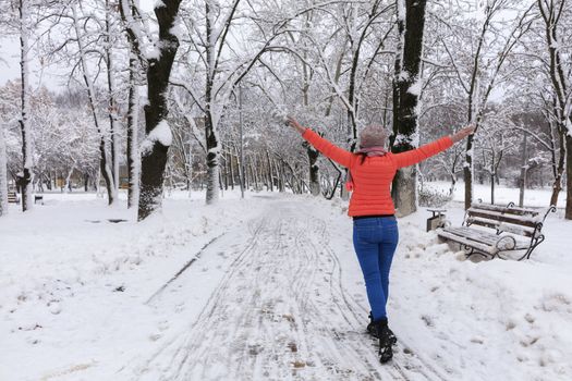 Young beautiful woman in a bright coral jacket and blue jeans in winter walks along the avenue of snow-covered fairy-tale park with wooden benches and opens her arms wide