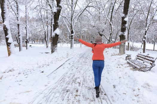 Young beautiful woman in a bright coral jacket and blue jeans in winter walks along the avenue of snow-covered fairy-tale park with wooden benches and opens her arms wide