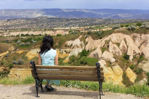 A young beautiful woman in a turquoise dress sits on a wooden bench on a hill and watches the panorama of the landscape of mountain caves and valleys in the canyons of Cappadocia
