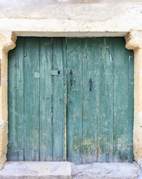 Very old weathered square wooden doors are painted with green paint with wrought handles and embedded in a wall made of shell rock.