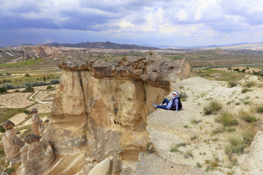 A young girl tourist with a backpack on her back is sitting on the edge of a cliff in Cappadocia and admiring the surrounding space against the background of a blue stormy sky and mountain scenery.