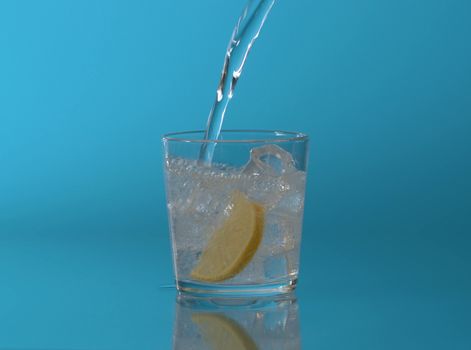 Close up tonic, soda sparkling water pouring into a glass with ice and slice of lemon on a blue background. Refreshing mineral water