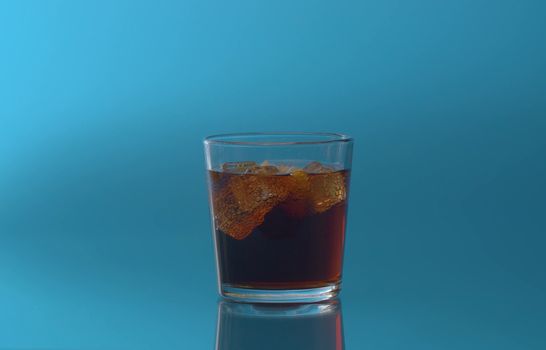 Glass of Cola with Ice and lemon close up. Cola fizzy drink with ice cubes and sliced lemon in transparent drinking glass over blue background. Coke Soda with bubbles closeup.
