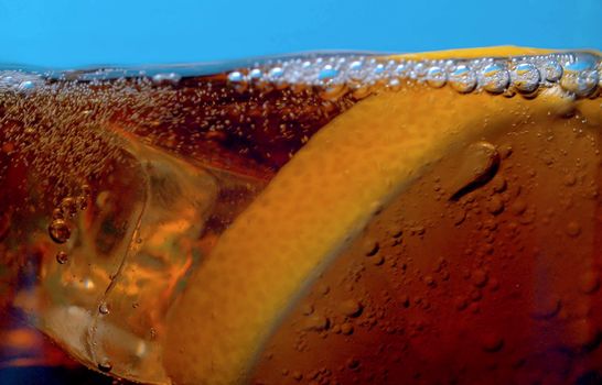 Rising gas bubbles in a glass with cola extreme close up. Sliced lemon and pieces of ice floating on the coca surface on blue background. Soda carbonated drink into a drinking glass