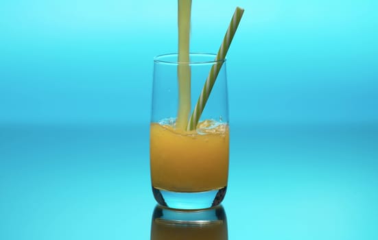 Fresh orange juice pouring into a transparent glass on a table. Close up refreshing yellow beverage on blue background. Healthy lifestyle concept