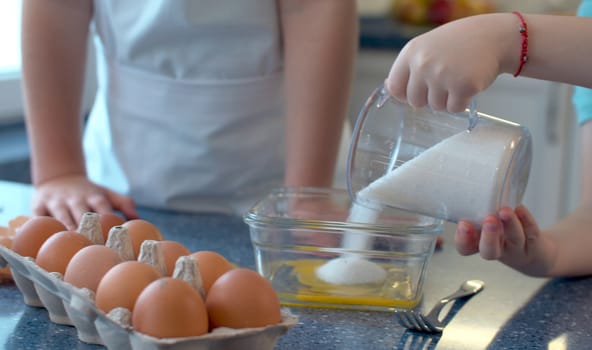 Close up child's hands pouring sugar into a glass bowl with broken eggs. Happy siblings are cooking pastries in a bright kitchen