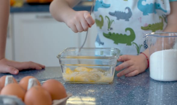 Close up child's hands pouring sugar into a glass bowl with broken eggs. Happy siblings are cooking pastries in a bright kitchen