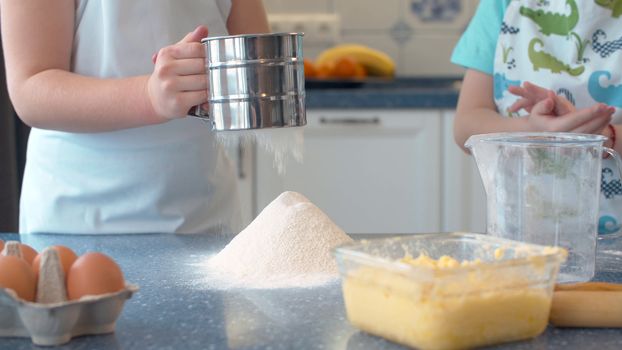 Close up kids cooking cakes in bright kitchen. Girl sifts flour through a sieve.