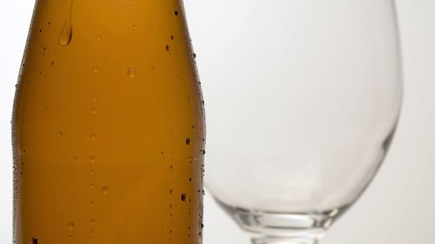 Close up misted bottle of cold beer on the table. Wineglass behind it. Macro shot on white background. Drops of water flowing down. Craft beer closeup