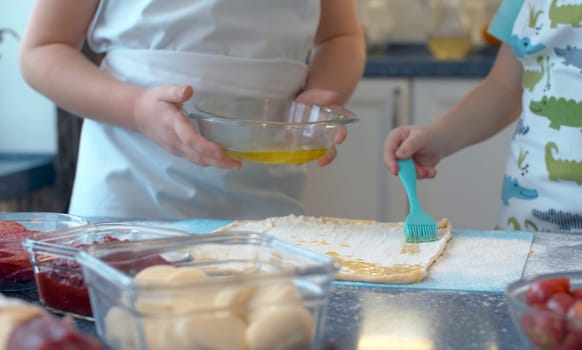 Close up hands of two young chefs cooking pizza. A girl holding a bowl with ghee and a boy covering a rolled dough with it, using a cooker brush.