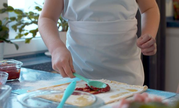 Close up hands young chefs cooking pizza. Girl putting tomato paste on rolled dough.