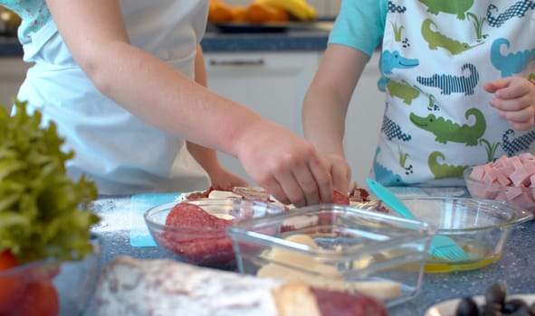 Close up hands of two children making pizza. Sister and brother putting sliced salami and ham on rolled dough. Slow motion