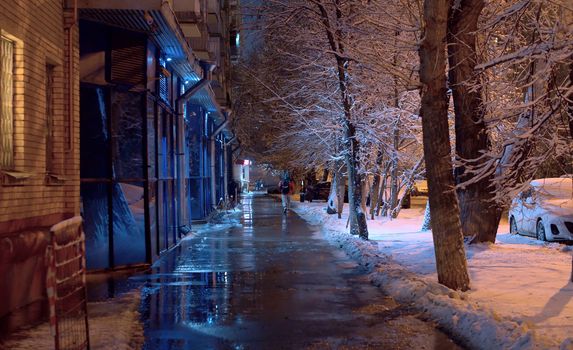 City street on a winter snowy night. The sidewalk is covered with water and ice, drops are falling from the roof. The light of lanterns is reflected in asphalt. Unrecognizable people