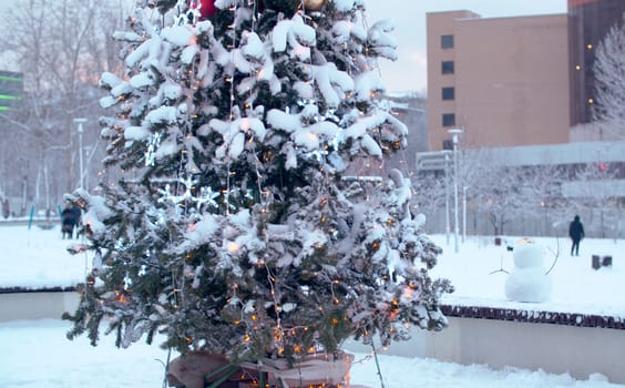 Christmas tree outdoors under the snow. Lights of a garland. City holiday decorations. Snowfall in the city.