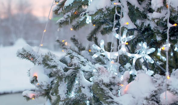 Close up snow covered branches of Christmas tree outdoors. Flickering lights of a garland. City holiday decorations. Snowfall in the city.