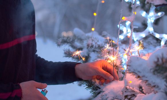 Close up male hand setting fire to sparklers on a Christmas tree. Blurry lights of a garland. City holiday decorations. Snowfall in the city.