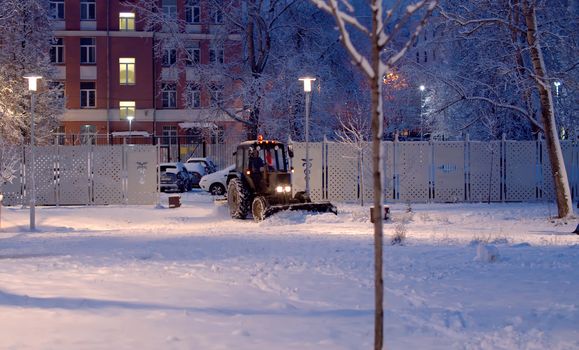 Tractor grader removing snow from the road in the snow covered park. Winter evening. Light of lanterns, luminous windows in the houses. Road service in work