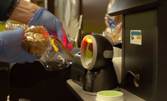 Close up hands in gloves scanning bread on a self-service scanner at a supermarket. Self-checkout counter in the food store during coronavirus epidemic.