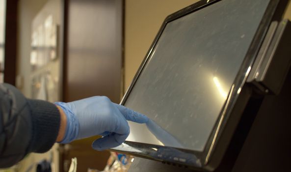 Close up hand of man at supermarket typing on touch screen of self checkout. Protective rubber blue gloves. Coronavirus epidemic in the city