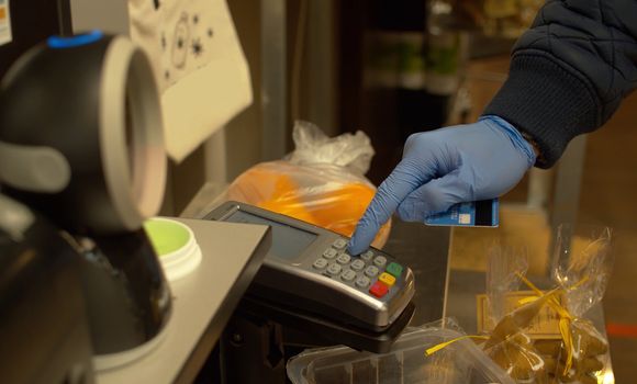 Close up hand of a man dialing pin code on pos terminal at self checkout counter inside foods store. Male hand in latex gloves paying by credit card during coronavirus epidemic