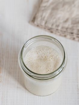 Wheat sourdough starter. Top view of glass jar with sourdough starter on white wooden background. Copy space for text or design. Vertical.