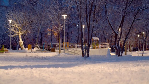 Snow covered city park in the evening. Light of lanterns. Winter cityscape. Children walking on the kid's playground
