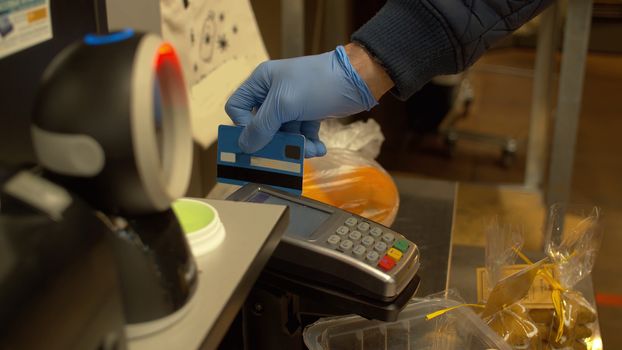 Close up hand of a man swiping debit card on pos terminal at self checkout counter inside foods store. Male hand in latex gloves paying by credit card during coronavirus epidemic