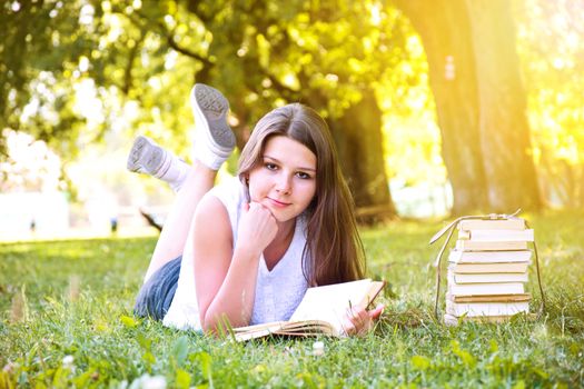 Young beautiful college student girl lying down on the green grass and reading a book at campus at warm day. Education. Back to school conceptual image.