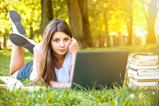 Young beautiful college student girl lying down on the green grass and working on laptop at campus at warm day. Education.