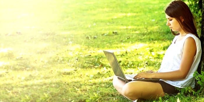 Young beautiful college student girl sitting on the green grass and working on laptop at campus at warm day. Education.