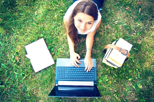 Young beautiful college student girl lying down on the green grass and working on laptop at campus. Education. Instagram vintage picture.