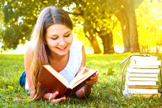 Young beautiful college student girl lying down on the green grass and reading a book at campus at warm day. Education. Back to school conceptual image.