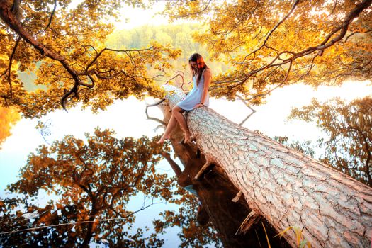 Autumn in the forest and lake. Beautiful girl sitting on the fallen tree over the water.