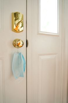 Facemask hanging from door handle on white door with sunlight coming in through window.