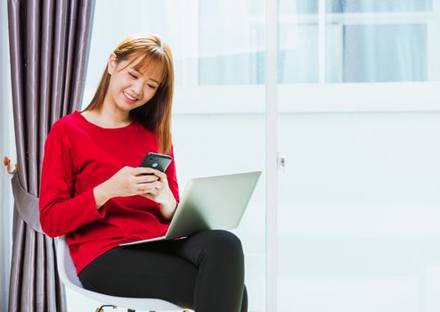 Work from home concept, Asian beautiful young woman smiling, student girl sitting on chair using mobile phone while typing or reading message text during working with laptop computer at home office